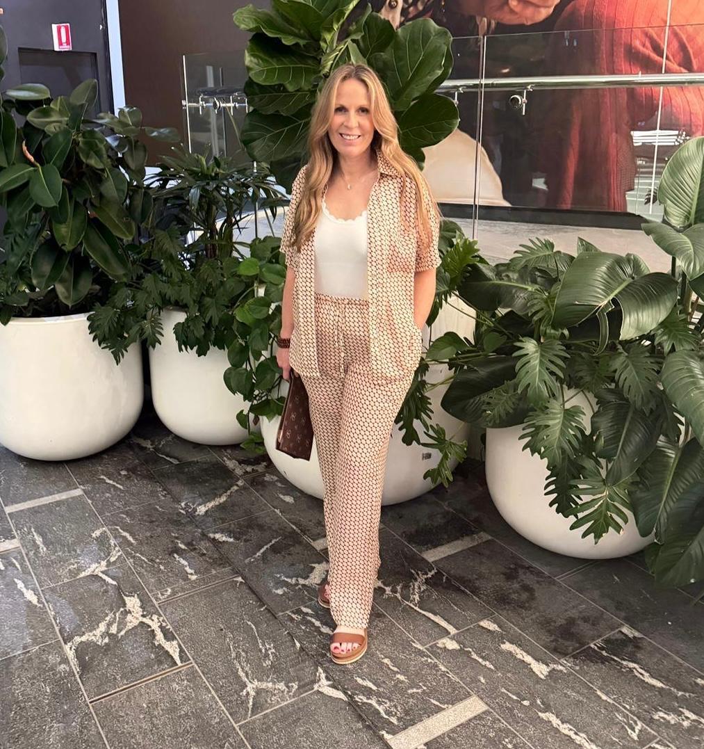 Ali standing in front of large green plants wearing a patterned shirt and pants